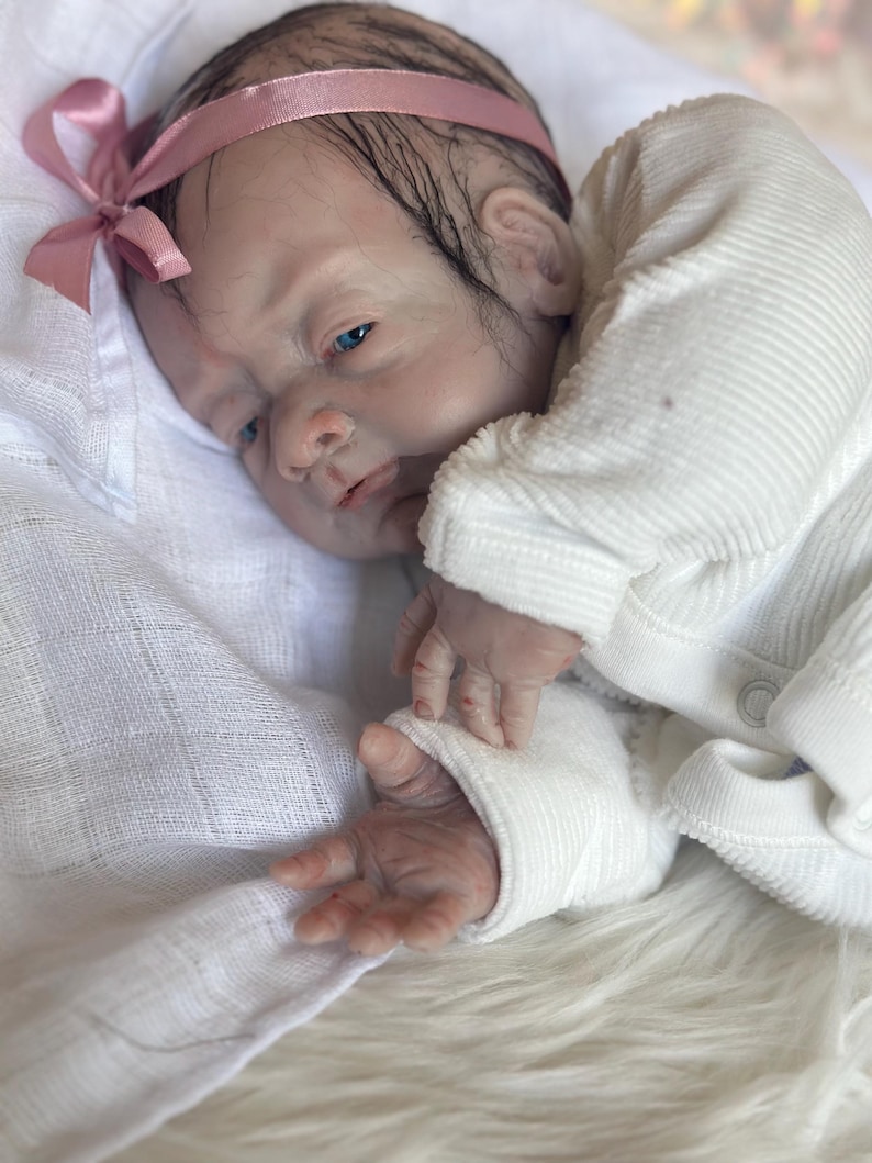 May include: A realistic reborn doll with pale skin, blue eyes, and dark hair, wearing a white long-sleeved ribbed shirt and a pink satin headband with a bow. The doll is resting on a white fabric surface, showcasing detailed facial features.