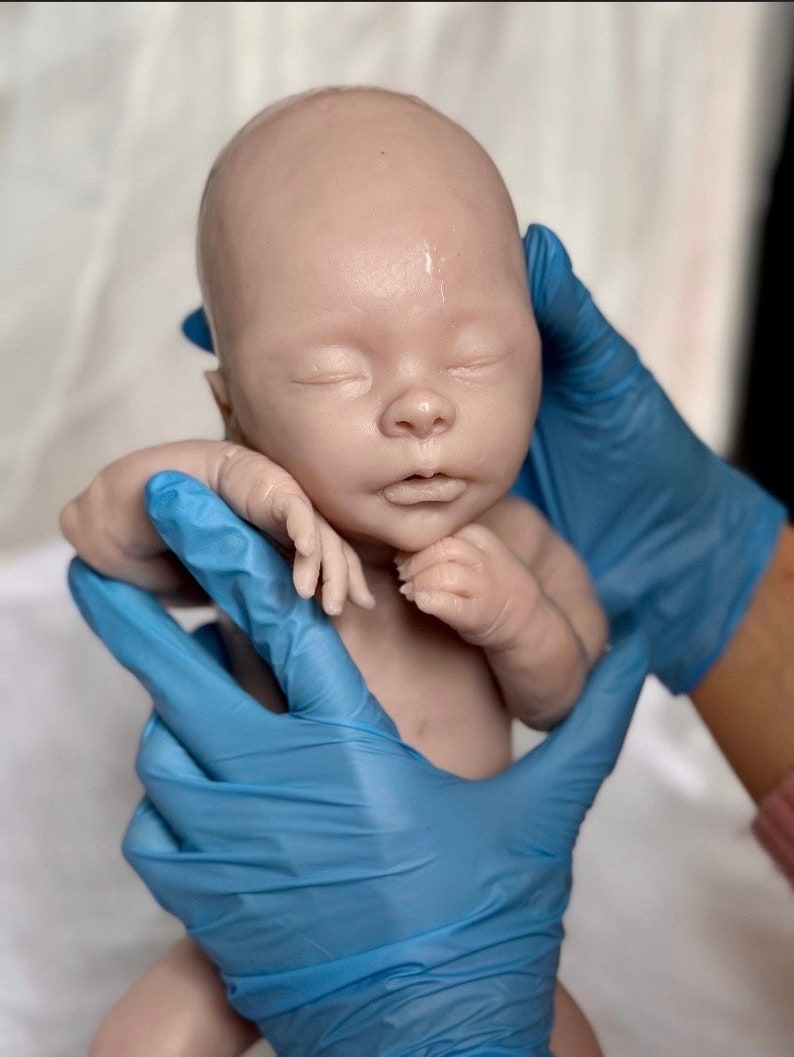 Puede incluir: Un mu&ntilde;eco beb&eacute; realista con los ojos cerrados y una tez rosa claro. El mu&ntilde;eco est&aacute; siendo sostenido por manos enguantadas.