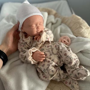 May include: A realistic baby doll with closed eyes, wearing a white pointed cap and a cream-colored long-sleeved footed sleeper featuring a repeating pattern of small brown deer and pink flowers, accompanied by a small round pink pacifier.