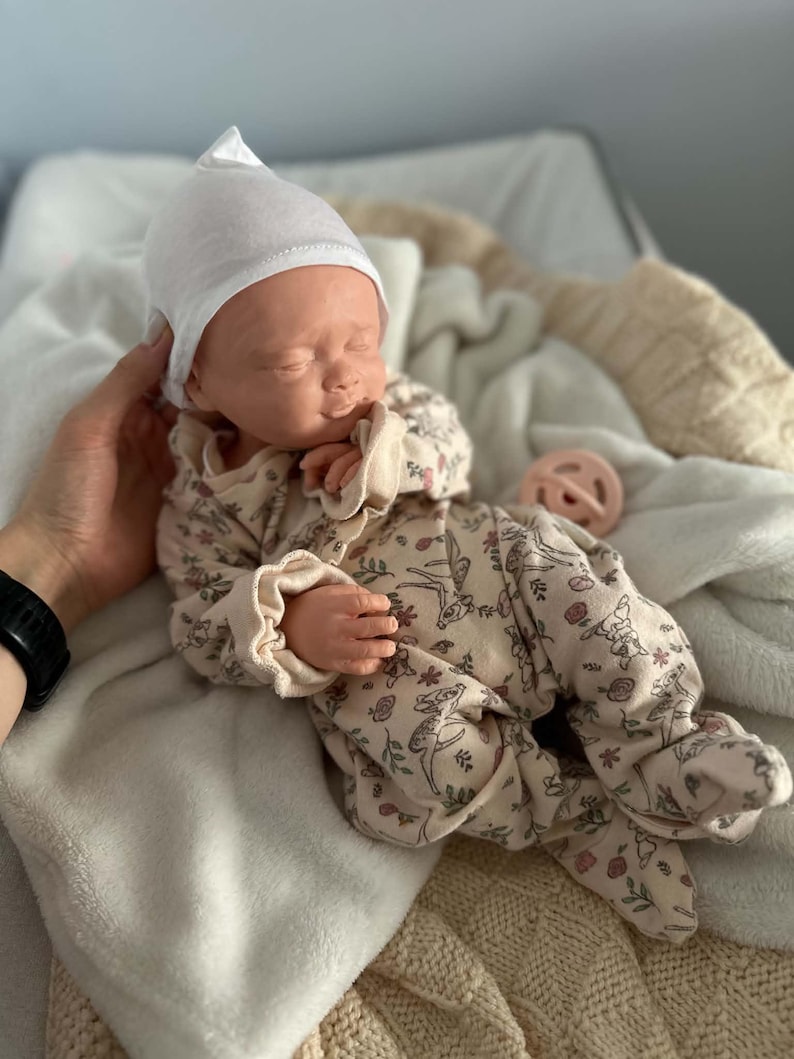 May include: A realistic baby doll with closed eyes, wearing a white pointed cap and a cream-colored long-sleeved footed sleeper featuring a repeating pattern of small brown deer and pink flowers, accompanied by a small round pink pacifier.