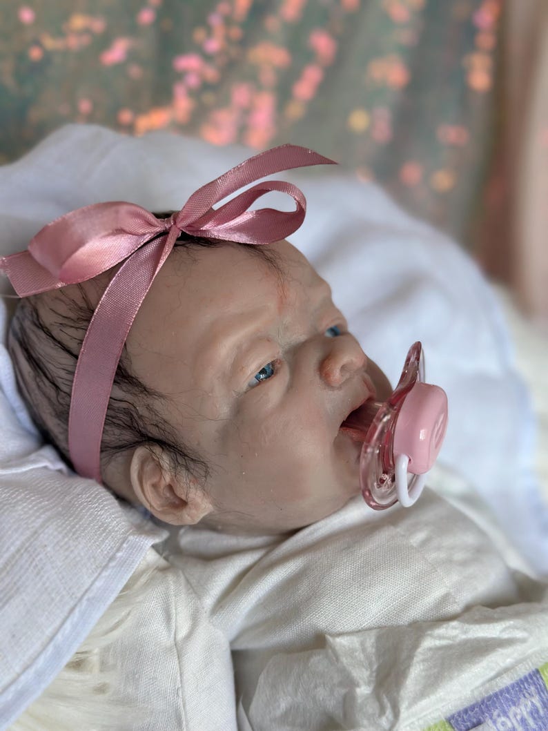 May include: A realistic reborn doll with fair skin, blue eyes, and sparse dark hair. The doll wears a pink satin ribbon headband and a white garment, with a clear and pink plastic pacifier positioned in its mouth, resting on a white fabric.