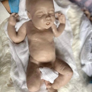 May include: A realistic baby doll with closed eyes and a slightly open mouth, lying on a white fluffy blanket. The doll is wearing a white cloth diaper and has a soft, lifelike appearance.