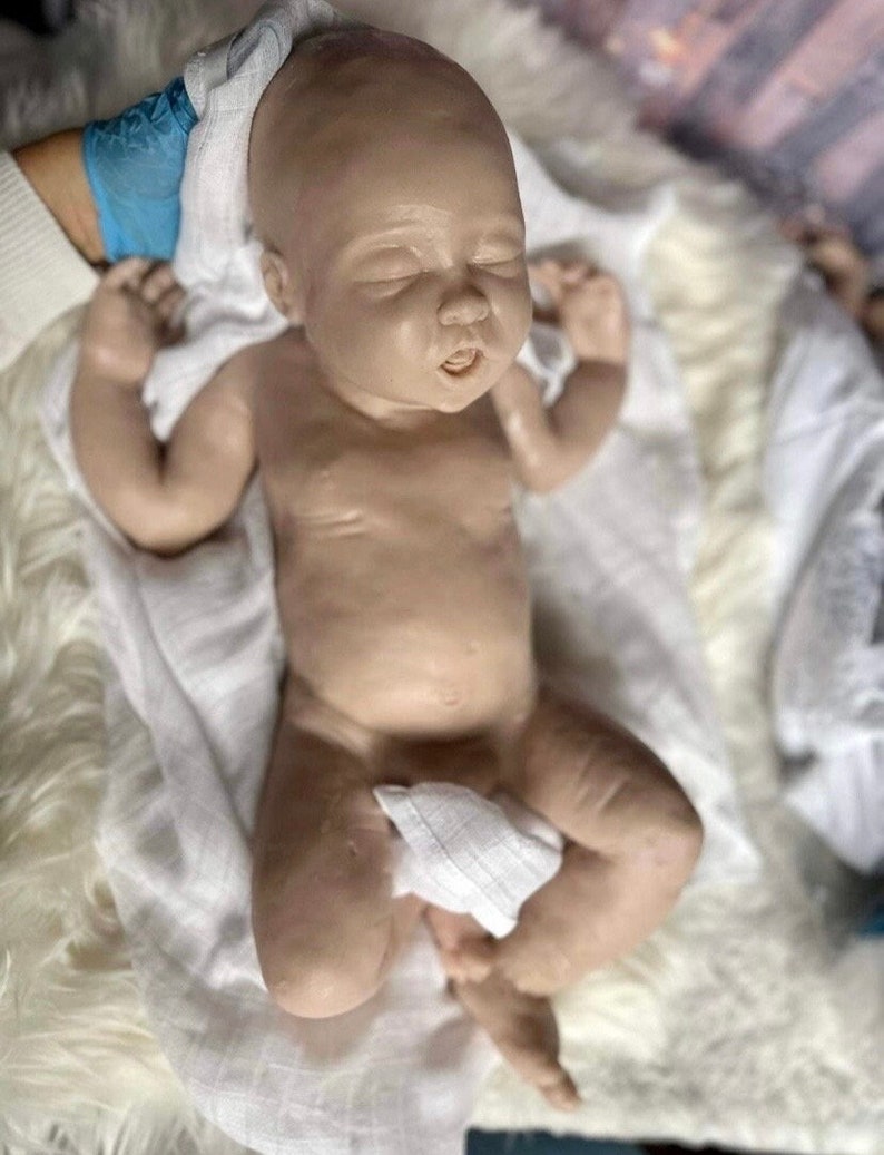 May include: A realistic baby doll with closed eyes and a slightly open mouth, lying on a white fluffy blanket. The doll is wearing a white cloth diaper and has a soft, lifelike appearance.
