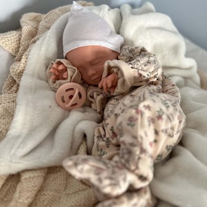 May include: A realistic baby doll with closed eyes, wearing a white knit cap and a cream-colored long-sleeved footed sleeper featuring a delicate floral and animal print pattern. A soft, round, pale pink pacifier rests near the doll's hand.