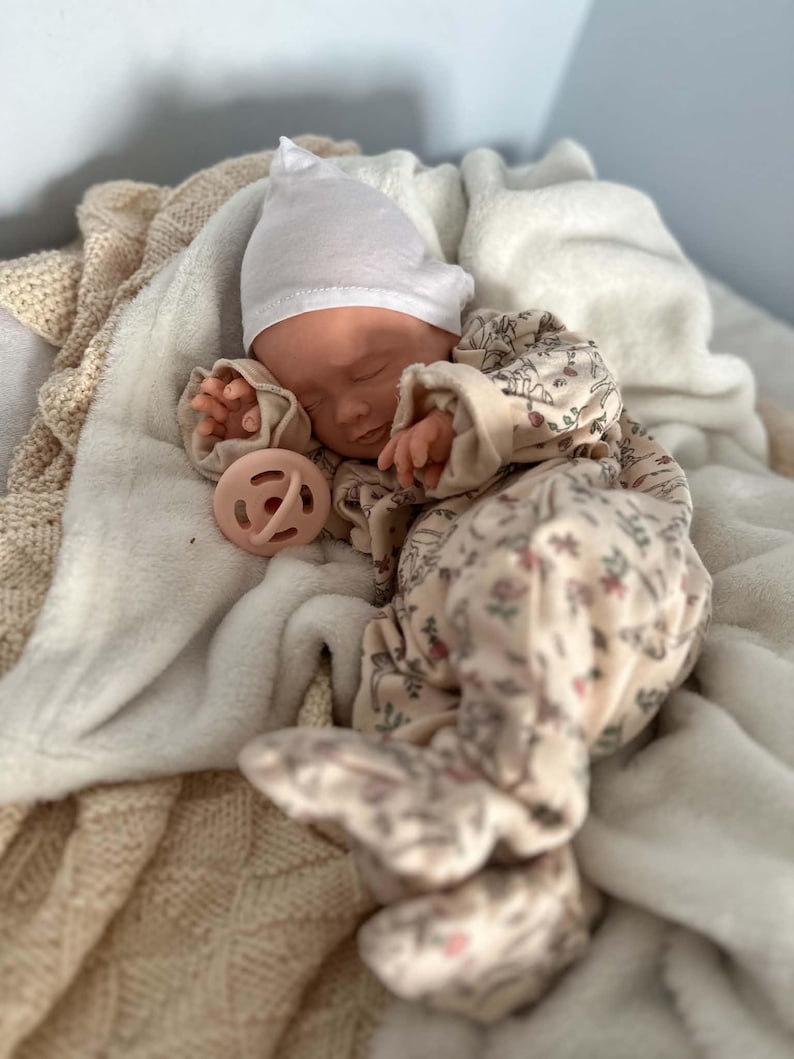 May include: A realistic baby doll with closed eyes, wearing a white knit cap and a cream-colored long-sleeved footed sleeper featuring a delicate floral and animal print pattern. A soft, round, pale pink pacifier rests near the doll's hand.