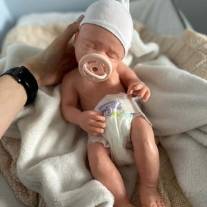 May include: A realistic newborn baby doll with light skin, closed eyes, and a soft white cap. The doll is wearing a white diaper with a colorful graphic print and has a light pink pacifier in its mouth. The doll is resting on a soft white blanket.