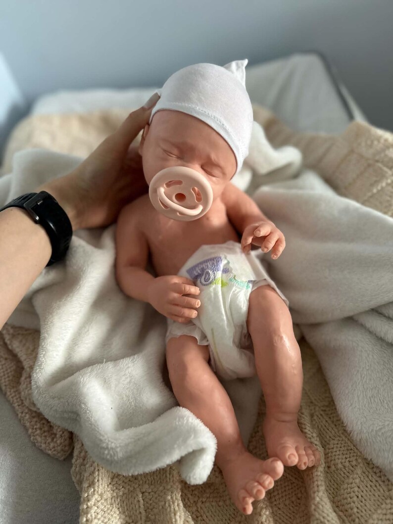 May include: A realistic newborn baby doll with light skin, closed eyes, and a soft white cap. The doll is wearing a white diaper with a colorful graphic print and has a light pink pacifier in its mouth. The doll is resting on a soft white blanket.