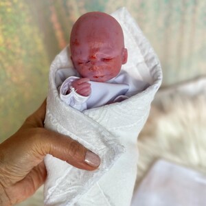 K&ouml;nnte beinhalten: Eine kleine, realistische Puppe mit rosiger Haut und geschlossenen Augen, eingewickelt in eine wei&szlig;e Wickeldecke mit einem dezenten, strukturierten Blumenmuster. Die Puppe tr&auml;gt ein schlichtes wei&szlig;es Langarmshirt unter der Decke.