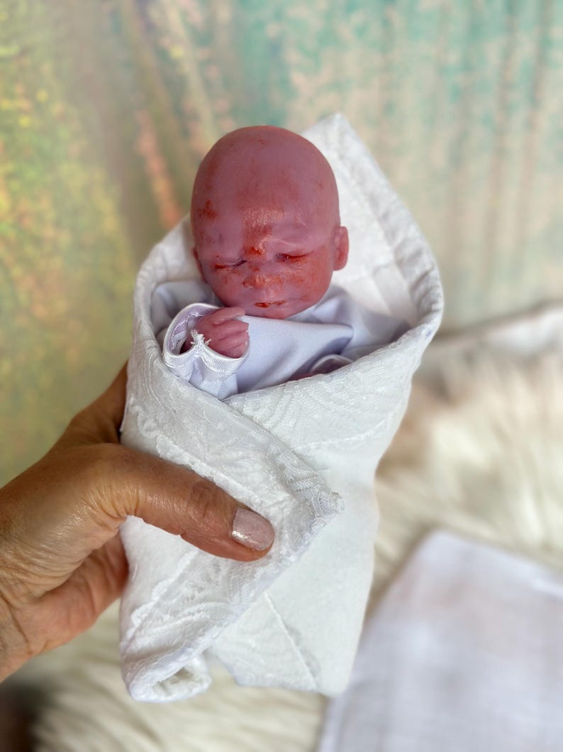 K&ouml;nnte beinhalten: Eine kleine, realistische Puppe mit rosiger Haut und geschlossenen Augen, eingewickelt in eine wei&szlig;e Wickeldecke mit einem dezenten, strukturierten Blumenmuster. Die Puppe tr&auml;gt ein schlichtes wei&szlig;es Langarmshirt unter der Decke.