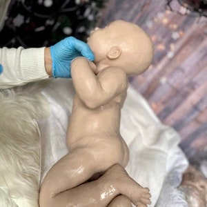 May include: A realistic silicone baby doll with a light brown complexion, lying on a white blanket. The doll has a slightly open mouth and closed eyes, and its arms and legs are bent.