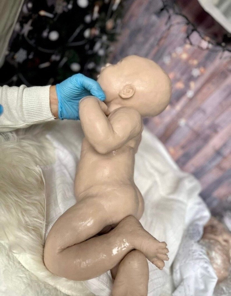 May include: A realistic silicone baby doll with a light brown complexion, lying on a white blanket. The doll has a slightly open mouth and closed eyes, and its arms and legs are bent.
