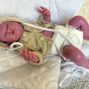 Puede incluir: Una mu&ntilde;eca beb&eacute; realista con un c&aacute;rdigan de punto beige y gorro a juego, adem&aacute;s de un pa&ntilde;al. La mu&ntilde;eca tiene un tono de piel realista y est&aacute; acostada sobre una manta blanca acolchada. El c&aacute;rdigan tiene botones y una capucha.