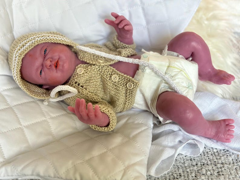 Puede incluir: Una mu&ntilde;eca beb&eacute; realista con un c&aacute;rdigan de punto beige y gorro a juego, adem&aacute;s de un pa&ntilde;al. La mu&ntilde;eca tiene un tono de piel realista y est&aacute; acostada sobre una manta blanca acolchada. El c&aacute;rdigan tiene botones y una capucha.