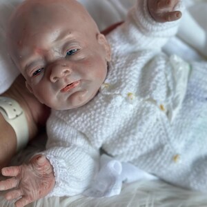 May include: A realistic reborn doll with pale skin, blue eyes, and subtle red mottling on the face and hands. The doll is wearing a white long-sleeved knitted cardigan with small yellow buttons and matching knitted pants, featuring a textured pattern.