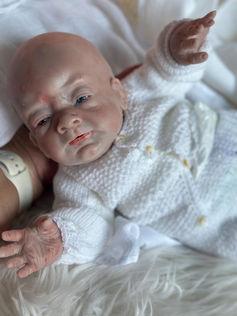 May include: A realistic reborn doll with pale skin, blue eyes, and subtle red mottling on the face and hands. The doll is wearing a white long-sleeved knitted cardigan with small yellow buttons and matching knitted pants, featuring a textured pattern.