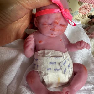 K&ouml;nnte beinhalten: Eine kleine, rosafarbene Silikonpuppe mit geschlossenen Augen, die eine wei&szlig;e Windel mit dem blauen Aufdruck Happy tr&auml;gt. Die Puppe hat ein leuchtend rosa Schleifenband um den Kopf und weist dezente rote Hautflecken im Gesicht und auf der Brust auf.