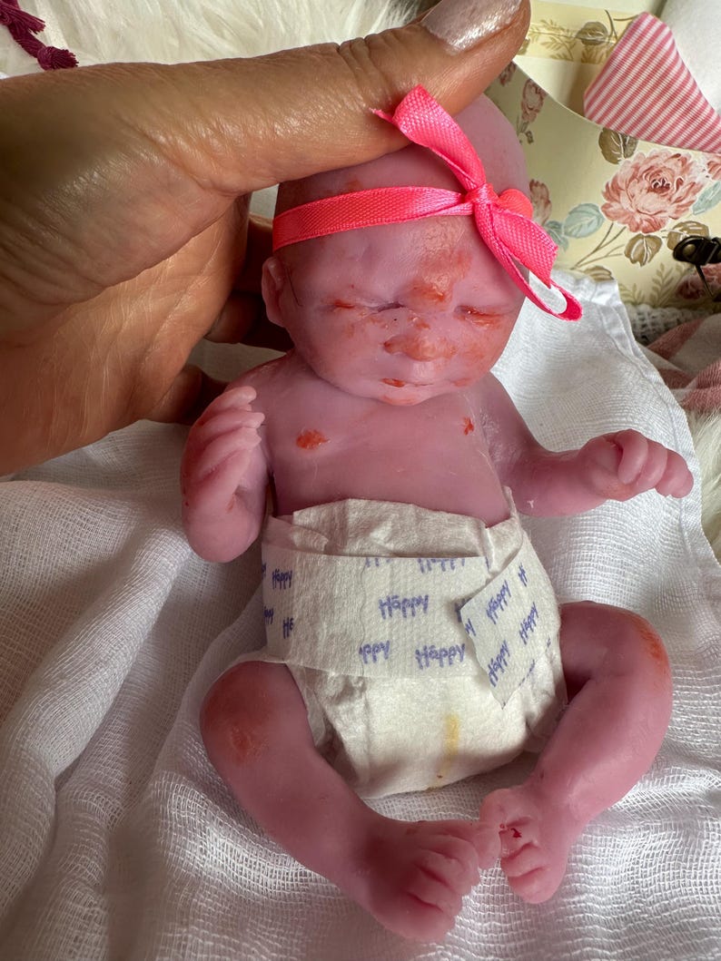 K&ouml;nnte beinhalten: Eine kleine, rosafarbene Silikonpuppe mit geschlossenen Augen, die eine wei&szlig;e Windel mit dem blauen Aufdruck Happy tr&auml;gt. Die Puppe hat ein leuchtend rosa Schleifenband um den Kopf und weist dezente rote Hautflecken im Gesicht und auf der Brust auf.