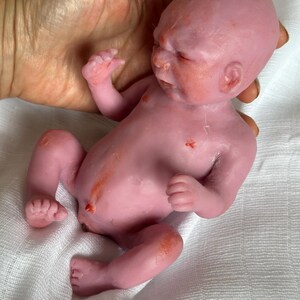 K&ouml;nnte beinhalten: Eine kleine, blassrosa Silikonpuppe, die einem Neugeborenen nachempfunden ist. Die Puppe hat eine realistische Hautstruktur, dezente rot-orangefarbene Hautt&ouml;ne, einen definierten Bauchnabel und geschlossene Augen in liegender Position.