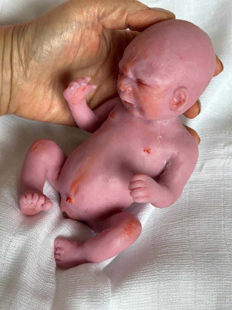 K&ouml;nnte beinhalten: Eine kleine, blassrosa Silikonpuppe, die einem Neugeborenen nachempfunden ist. Die Puppe hat eine realistische Hautstruktur, dezente rot-orangefarbene Hautt&ouml;ne, einen definierten Bauchnabel und geschlossene Augen in liegender Position.