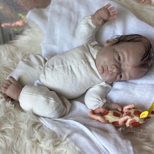 May include: A realistic reborn doll with painted hair and blue eyes, wearing a long-sleeved cream-colored onesie. The doll is lying on its back with one arm raised and is holding a small, plush giraffe toy with brown spots and yellow ears.