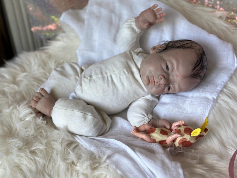 May include: A realistic reborn doll with painted hair and blue eyes, wearing a long-sleeved cream-colored onesie. The doll is lying on its back with one arm raised and is holding a small, plush giraffe toy with brown spots and yellow ears.