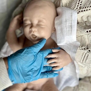 May include: A realistic baby doll with closed eyes and an open mouth, lying on a white blanket with a white cloth over its head. A gloved hand is touching the doll's hand.