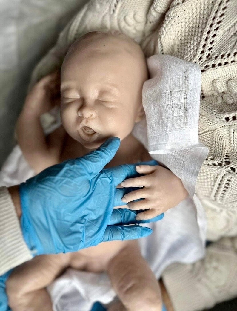 May include: A realistic baby doll with closed eyes and an open mouth, lying on a white blanket with a white cloth over its head. A gloved hand is touching the doll's hand.