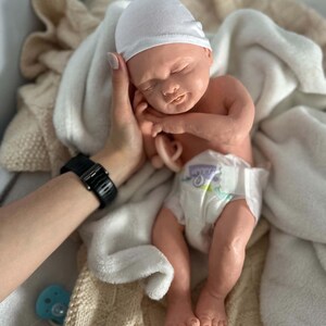May include: A realistic newborn baby doll with light skin, closed eyes, and a peaceful expression. The doll wears a plain white beanie cap and a disposable diaper with a printed pattern. A small, light-colored pacifier is held near the doll's chest.