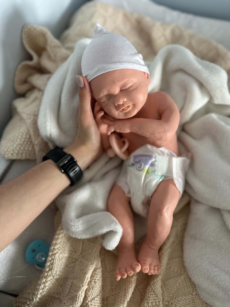 May include: A realistic newborn baby doll with light skin, closed eyes, and a peaceful expression. The doll wears a plain white beanie cap and a disposable diaper with a printed pattern. A small, light-colored pacifier is held near the doll's chest.