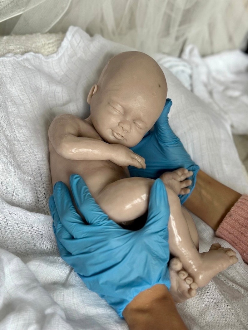 K&ouml;nnte beinhalten: Eine realistische, schlafende Babypuppe mit hellem Hautton wird von H&auml;nden mit blauen Handschuhen gehalten. Die Puppe liegt auf einem wei&szlig;en Tuch. Die Augen des Babys sind geschlossen, und seine Arme und Beine sind sanft gekr&uuml;mmt.