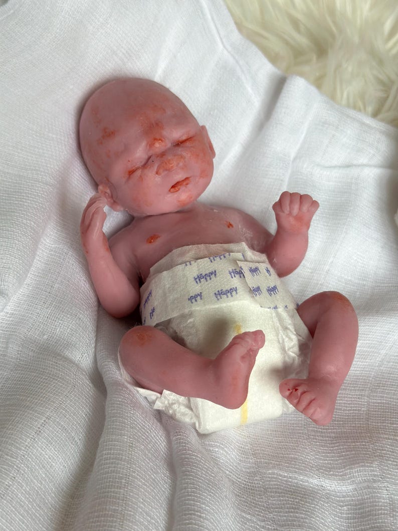 K&ouml;nnte beinhalten: Eine kleine, realistische Silikonpuppe mit rosigem Hautton und dezenter roter Sprenkelung, mit geschlossenen Augen und neutralem Ausdruck. Die Puppe tr&auml;gt eine wei&szlig;e Einwegwindel mit dem Wort Happy in blauem Aufdruck auf der Vorderseite.
