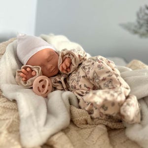 May include: A realistic baby doll with closed eyes, wearing a plain white beanie and a cream-colored long-sleeved onesie featuring a delicate floral and animal print. The doll is positioned with a solid light pink pacifier resting near its chest.