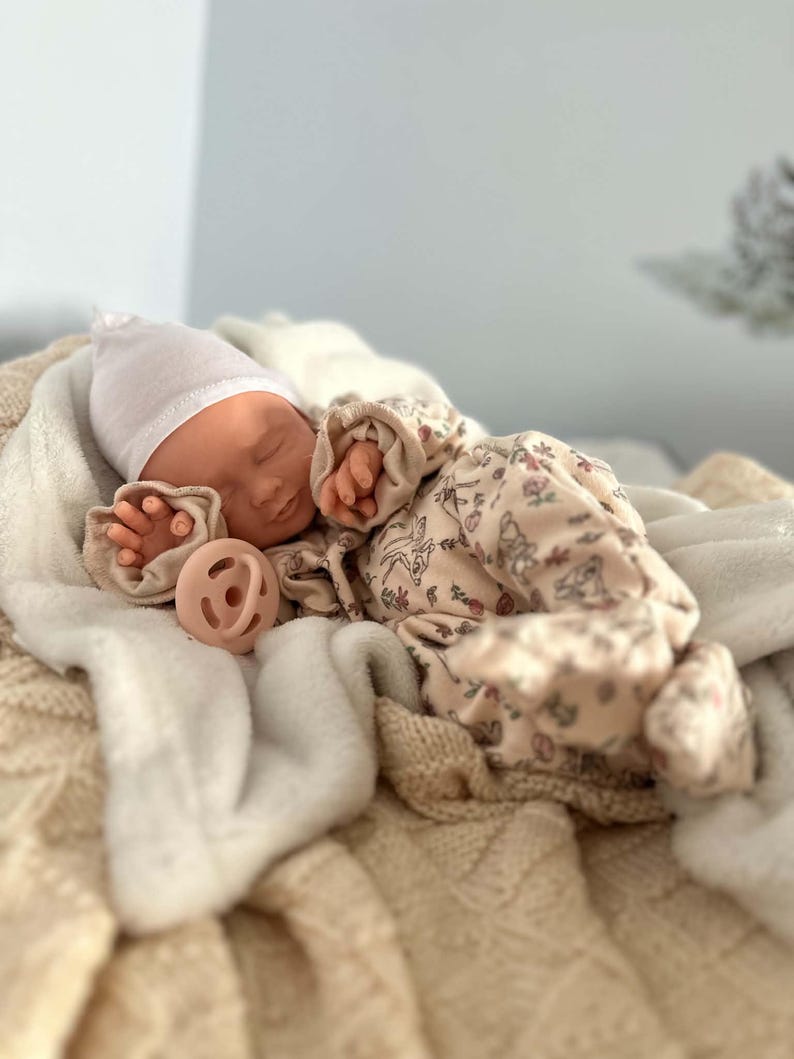 May include: A realistic baby doll with closed eyes, wearing a plain white beanie and a cream-colored long-sleeved onesie featuring a delicate floral and animal print. The doll is positioned with a solid light pink pacifier resting near its chest.