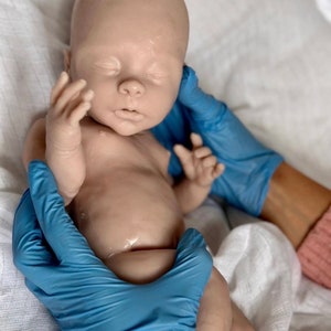 Puede incluir: Un mu&ntilde;eco de beb&eacute; de silicona realista con los ojos cerrados, acostado sobre una superficie de tela blanca. El mu&ntilde;eco est&aacute; siendo sostenido por dos manos enguantadas.
