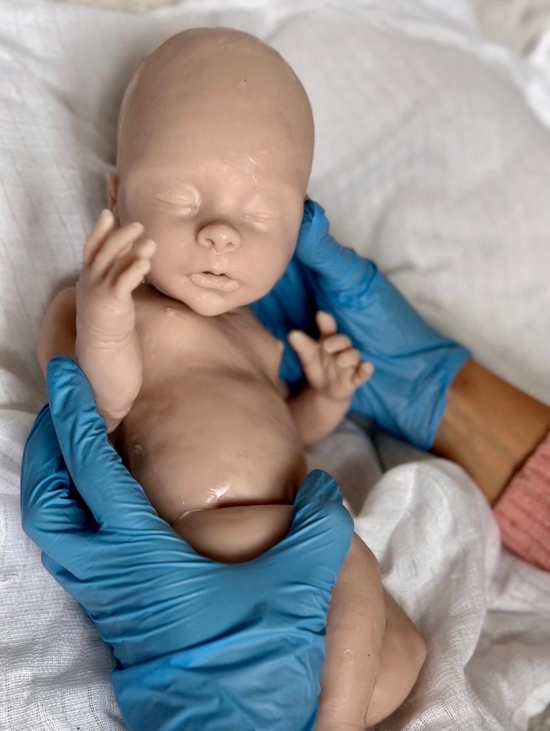 Puede incluir: Un mu&ntilde;eco de beb&eacute; de silicona realista con los ojos cerrados, acostado sobre una superficie de tela blanca. El mu&ntilde;eco est&aacute; siendo sostenido por dos manos enguantadas.