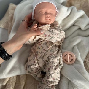 May include: A realistic newborn baby doll with closed eyes, wearing a white beanie and a beige footed sleeper featuring a repeating pattern of small deer, flowers, and foliage. A small, round, light pink pacifier is positioned next to the doll.
