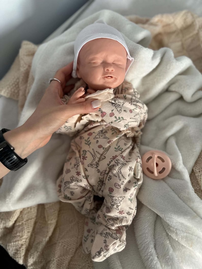 May include: A realistic newborn baby doll with closed eyes, wearing a white beanie and a beige footed sleeper featuring a repeating pattern of small deer, flowers, and foliage. A small, round, light pink pacifier is positioned next to the doll.