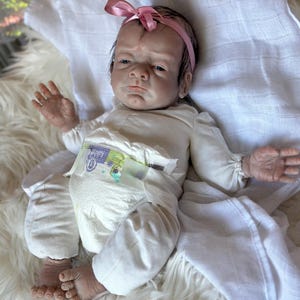 May include: A realistic newborn doll with dark hair and a pink ribbon headband, wearing a long-sleeved cream-colored shirt and a disposable diaper with a green and purple truck graphic print, lying on a white fabric surface.