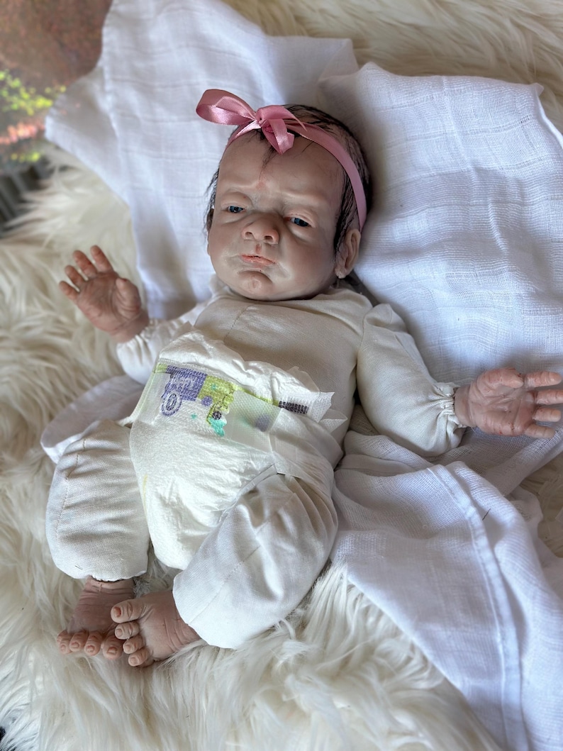 May include: A realistic newborn doll with dark hair and a pink ribbon headband, wearing a long-sleeved cream-colored shirt and a disposable diaper with a green and purple truck graphic print, lying on a white fabric surface.