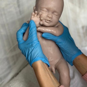 K&ouml;nnte beinhalten: Eine realistische Babypuppe in neutralem Farbton mit geschlossenen Augen und friedlichem Ausdruck. Die Puppe wird von H&auml;nden mit blauen Handschuhen gehalten. Die Haut des Babys wirkt glatt, mit detaillierten Merkmalen. Der Gesamteindruck ist der eines Neugeborenen.
