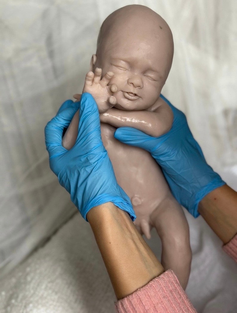 K&ouml;nnte beinhalten: Eine realistische Babypuppe in neutralem Farbton mit geschlossenen Augen und friedlichem Ausdruck. Die Puppe wird von H&auml;nden mit blauen Handschuhen gehalten. Die Haut des Babys wirkt glatt, mit detaillierten Merkmalen. Der Gesamteindruck ist der eines Neugeborenen.