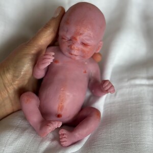 K&ouml;nnte beinhalten: Eine Miniatur-Neugeborenenpuppe mit blassrosa Haut, die eine dezente r&ouml;tlich-orangefarbene Sprenkelung im Gesicht, auf der Brust und am Bauch aufweist. Die Puppe hat geschlossene Augen, eine kleine Nase und einen definierten Bauchnabel.