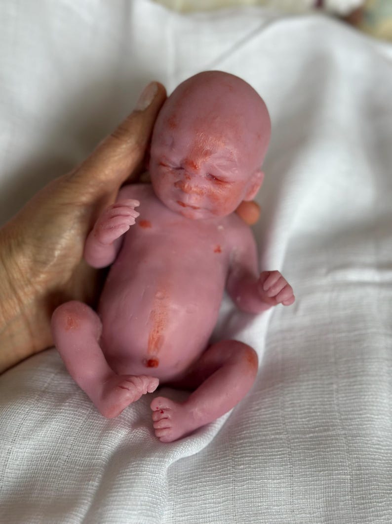 K&ouml;nnte beinhalten: Eine Miniatur-Neugeborenenpuppe mit blassrosa Haut, die eine dezente r&ouml;tlich-orangefarbene Sprenkelung im Gesicht, auf der Brust und am Bauch aufweist. Die Puppe hat geschlossene Augen, eine kleine Nase und einen definierten Bauchnabel.