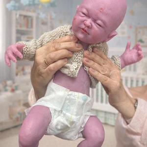 Puede incluir: Una mu&ntilde;eca beb&eacute; realista, de tono p&uacute;rpura, con un c&aacute;rdigan beige tejido y un pa&ntilde;al. La mu&ntilde;eca tiene una cara realista con los ojos cerrados y un ligero ce&ntilde;o fruncido. La mu&ntilde;eca est&aacute; siendo sostenida por una persona.