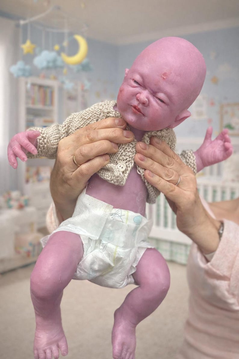 Puede incluir: Una mu&ntilde;eca beb&eacute; realista, de tono p&uacute;rpura, con un c&aacute;rdigan beige tejido y un pa&ntilde;al. La mu&ntilde;eca tiene una cara realista con los ojos cerrados y un ligero ce&ntilde;o fruncido. La mu&ntilde;eca est&aacute; siendo sostenida por una persona.