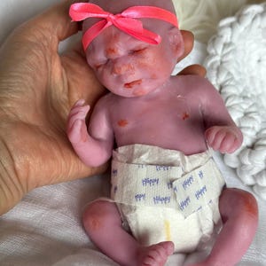 K&ouml;nnte beinhalten: Eine kleine, realistische Silikonpuppe mit violettem Teint, die ein leuchtend neonpinkes Band um den Kopf tr&auml;gt. Die Puppe tr&auml;gt eine wei&szlig;e Einwegwindel, auf deren Vorderseite das Wort Happy in blauer Schrift aufgedruckt ist.