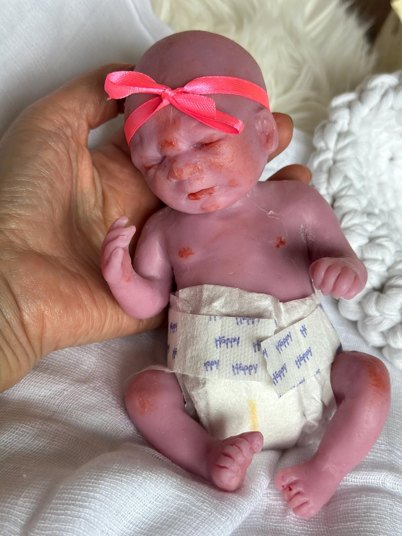 K&ouml;nnte beinhalten: Eine kleine, realistische Silikonpuppe mit violettem Teint, die ein leuchtend neonpinkes Band um den Kopf tr&auml;gt. Die Puppe tr&auml;gt eine wei&szlig;e Einwegwindel, auf deren Vorderseite das Wort Happy in blauer Schrift aufgedruckt ist.