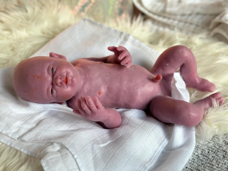 Puede incluir: Una mu&ntilde;eca beb&eacute; realista, de tono malva, yace sobre una tela blanca, sobre un fondo esponjoso blanquecino. La mu&ntilde;eca tiene rasgos detallados, incluyendo una frente arrugada y peque&ntilde;as manos y pies. La mu&ntilde;eca beb&eacute; es una representaci&oacute;n realista de un reci&eacute;n nacido.