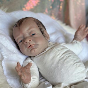 May include: A realistic reborn baby doll with light skin, painted dark hair, and bright blue eyes. The doll is wearing a long-sleeved, cream-colored onesie and is positioned with arms slightly raised, showcasing detailed facial features and skin texture.