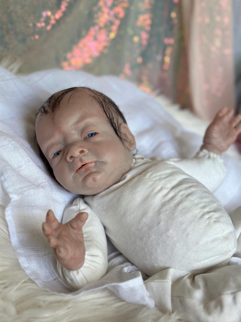 May include: A realistic reborn baby doll with light skin, painted dark hair, and bright blue eyes. The doll is wearing a long-sleeved, cream-colored onesie and is positioned with arms slightly raised, showcasing detailed facial features and skin texture.